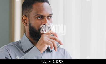 Lächelnder, fröhlicher junger afroamerikanischer Kerl, Mitarbeiter des Unternehmens hält Glas und trinkt reines kaltes Wasser. Gesunde Mischrasse bärtigen Mann Hydrierung Stockfoto