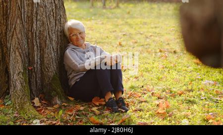 Ältere kaukasische Rentnerin verträumte Frau, die unter einem Baum sitzt, sich entspannt und sonniges Herbstwetter genießt. Außenaufnahme. Hochwertige Fotos Stockfoto