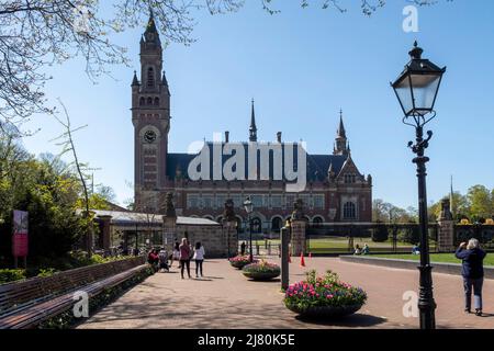 Touristen spazieren und machen Fotos in der Nähe des Friedenspalastes an einem sonnigen Tag in den Haag, Niederlande, umgeben von Frühlingsblumen und Bänken Stockfoto