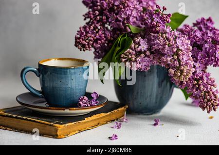 Tasse Kaffee auf einem Buch neben einer Vase mit Fliederblumen Stockfoto