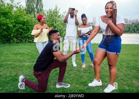 amerikanischer Mann, der auf einem Knie steht und Frau um Heirat bittet und einen Ring schlägt, überrascht schockiert, als er Gruppenfreunde überall in Schock versammelt Stockfoto