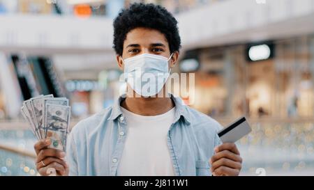 afroamerikanischer Mann Kerl männlichen Gewinner trägt medizinische Gesichtsmaske hält Bankkarte und Dollar Geld in den Händen macht die Wahl zwischen Bargeld und elektronisch Stockfoto