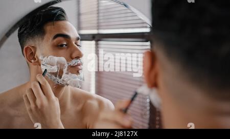 Hispanic araber indian bärtig schöner Mann Rasieren mit Einweg-Rasiermesser im Bad Blick auf Spiegel. Tägliche morgendliche Routine männlich hygienisch Stockfoto