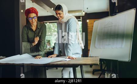 Fröhliche Architektinnen, die in einem modernen Büro an Blaupause-Zeichnungen arbeiten. Zwei muslimische Geschäftsfrauen planen ein neues innovatives Projekt. Kreatives Design Stockfoto