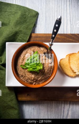 Hühnerleber-Mousse-Portion mit ein paar Stücken getrocknetem Brot, Knäckebrot serviert. Serviert in einer runden braunen Schüssel und mit einem Löffel in eine Schüssel getaucht. Dez Stockfoto