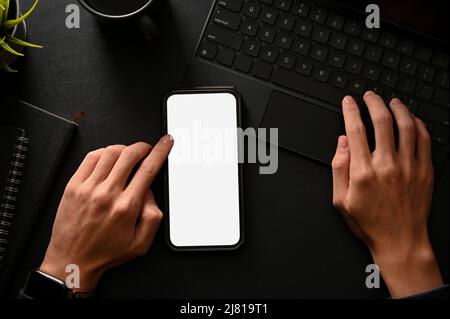 Draufsicht, flaches Lay, Ein Mann mit Smartphone und tippen auf der kabellosen Tablet-Tastatur in schwarzem Tischhintergrund. Modell mit weißem Bildschirm des Telefons. Stockfoto