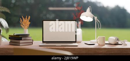 Komfortabler Arbeitsplatz mit leerem Laptop-Bildschirm, Tischlampe, Bücherstapel und Zubehör auf dem Tisch über verschwommenem grünen Feld im Hintergrund. Stockfoto