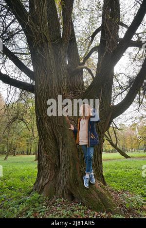 Das verträumte Teenager-Mädchen im Stadtpark steht im Frühherbst in der Nähe des riesigen Weidenbaums Stockfoto