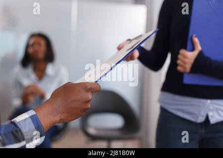 Nahaufnahme von Mann Hände geben HR-Abteilungsleiter Lebenslauf und abgeschlossene Bewerbung, während im Wartezimmer des Start-up-Unternehmen sitzen. Verschiedene Menschen im Lobbybereich der Marketingagentur. Stockfoto