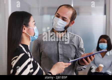 Positiver und selbstbewusster Arbeitgeber trägt eine Schutzmaske, während er mit dem Bewerber über den Lebenslauf spricht. Arbeitslose asiatische Frau, die im Lobbybereich Personalabteilung-Manager rekrutiert. Stockfoto