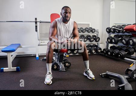 Ein afroamerikanischer Mann sitzt auf einer Bank im Fitnessstudio und ruht sich aus Stockfoto