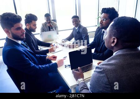 Chef-Afro-Geschäftsleute im Gespräch mit arabischen und indischen Partner Kollegen, multiethnische Team sitzt an einem Tisch mit einem Laptop und Dokumente im Büro Stockfoto