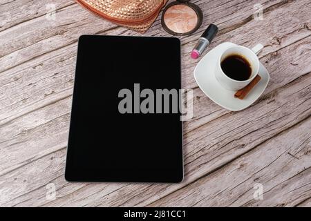 Tablet auf dem Tisch, umgeben von Make-up-Beutel, Lippenstift und Kaffeetasse. Stockfoto