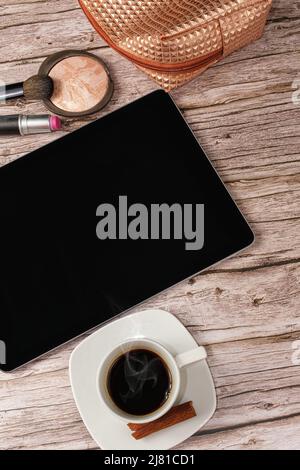 Tablet auf dem Tisch, umgeben von Make-up-Beutel, Lippenstift und Kaffeetasse vertikales Foto. Stockfoto