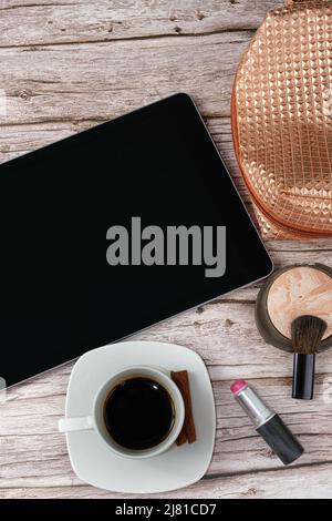 Tablet auf dem Tisch, umgeben von Make-up-Beutel, Lippenstift und Kaffeebecher Draufsicht. Stockfoto