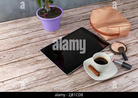 Tablet auf dem Tisch, umgeben von Kaffeetasse, Lippenstift und Make-up-Beutel. Stockfoto