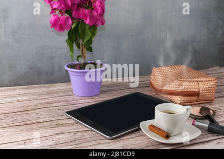 Tablet auf dem Tisch, umgeben von Kaffeetasse, Lippenstift und Make-up Bag Seitenansicht. Stockfoto