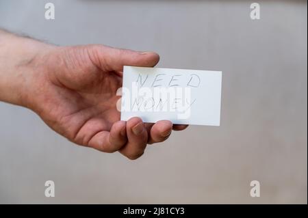 Brauchen Sie Geld. Wörter in gezackten Buchstaben. Die Hand eines Mannes hält ein weißes Papierrechteck mit Text vor einem grauen Hintergrund. Stockfoto