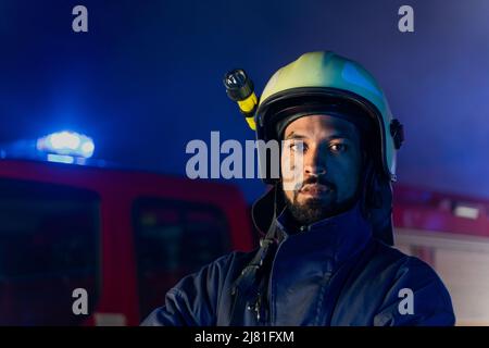 Porträt eines schmutzigen Feuerwehrmanns im Dienst mit einem Feuerwehrauto im Hintergrund in der Nacht, lächelnd. Stockfoto