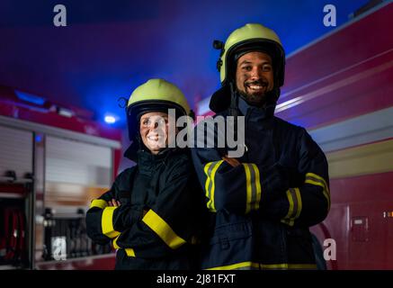 Glückliche Feuerwehrleute Mann und Frau nach der Aktion Blick auf Kamera mit Feuerwehrauto im Hintergrund Stockfoto