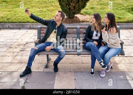 Fröhlich lächelnde Freunde im Park sitzen auf einer Bank und machen Selfies mit einem Smartphone. Hochwertige Fotos Stockfoto