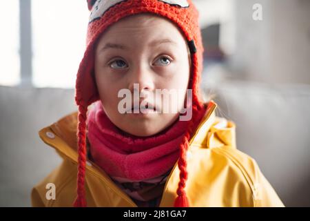 Kleines Mädchen mit Down-Syndrom, das im Winter die Kamera-Outoors gegen die Ziegelmauer anschaut. Stockfoto