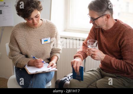 Junge Frau hilft dem ukrainischen Mann ehrenamtlich beim Ausfüllen von Formularen im Asylzentrum. Stockfoto