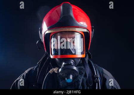 Portrait starker Feuerwehrmann in feuerfester Uniform, der eine Axt-Kettensäge in seinen Händen hält, schwarzes Hintergrundstudio.Sauerstoffmaske auf dem Kopf aus nächster Nähe Stockfoto