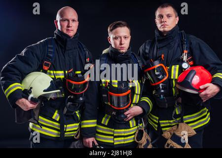 Portrait еркуу starker Feuerwehrmann in feuerfester Uniform mit Axt-Kettensäge in den Händen schwarzer Hintergrund Studio.Team Work Konzept Stockfoto