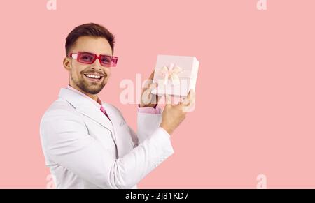 Glücklich attraktiven jungen Mann in Anzug zeigt Geschenkbox isoliert auf pastellrosa Hintergrund. Stockfoto