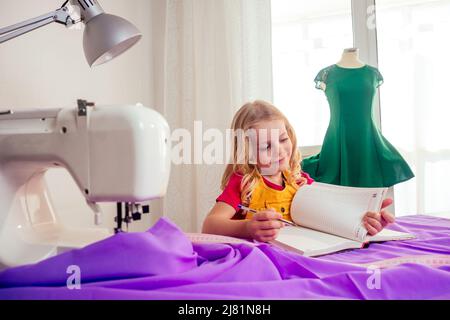 Lächelndes kleines blondes Mädchen am Tisch mit Nähmaschine mit Übungspuppe Stockfoto