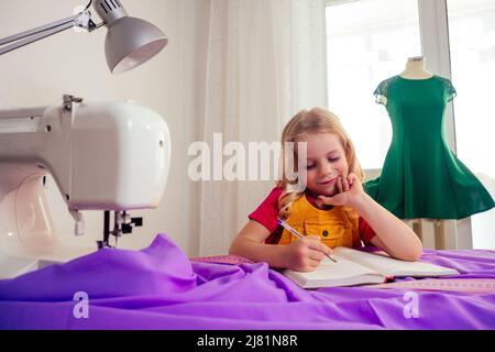 Lächelndes kleines blondes Mädchen am Tisch mit Nähmaschine mit Übungspuppe Stockfoto