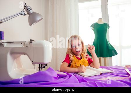 Lächelndes kleines blondes Mädchen am Tisch mit Nähmaschine mit Übungspuppe Stockfoto