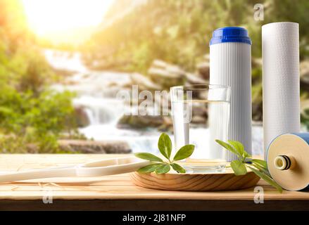 Glas Wasser gereinigt durch Osmose mit umweltfreundlichen biologisch abbaubaren Filtern auf Holzlatten Tisch mit Natur Hintergrund Bäume und Wasserfall Stockfoto