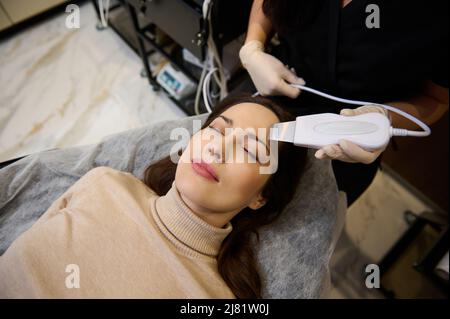 Nahaufnahme einer charmanten, schönen dunkelhaarigen kaukasischen Frau mittleren Alters, die im Wellness-Spa-Center ein Gesichtspeeling mit Ultraschallkavitation erhält. Haut c Stockfoto