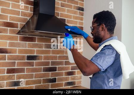 afrikanischer Mann, der die Dunstabzugshaube mit Kochplatte zu Hause putzt, Backsteinwand im Hintergrund Stockfoto