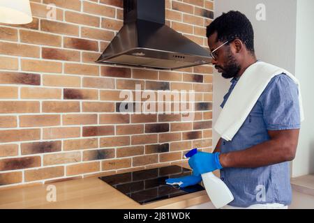 afrikanischer Mann, der die Dunstabzugshaube mit Kochplatte zu Hause putzt, Backsteinwand im Hintergrund Stockfoto