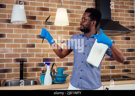 afrikanischer Mann, der die Dunstabzugshaube putzt und zu Hause wie ein Mikrofon einen Kochlöffel ansingt, Backsteinwand-Hintergrund Stockfoto