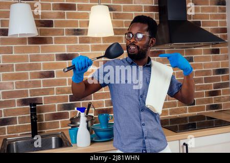 afrikanischer Mann, der die Dunstabzugshaube putzt und zu Hause wie ein Mikrofon einen Kochlöffel ansingt, Backsteinwand-Hintergrund Stockfoto