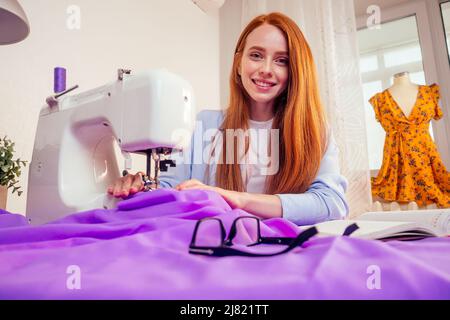 Rothaarige Geschäftsfrau mit Nähmaschine und maniken im Studio-Hintergrund. Schneider schafft eine Kollektion Outfits näht Kleidung in Werkstatt schreiben Stockfoto