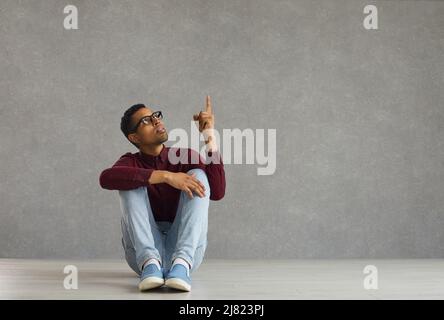 Inspirierter, dunkelhäutiger junger Mann mit erhobenem Finger sinniert auf grauem Hintergrund. Stockfoto