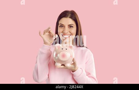 Lächelnde, sparsame Frau wirft eine Münze in ein Sparschwein, das sie auf rosa Hintergrund in der Hand hält. Stockfoto