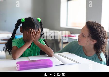 Kaukasische junge Lehrerin, die afroamerikanische Grundschülerin mit der Hand auf dem Gesicht anschaut Stockfoto