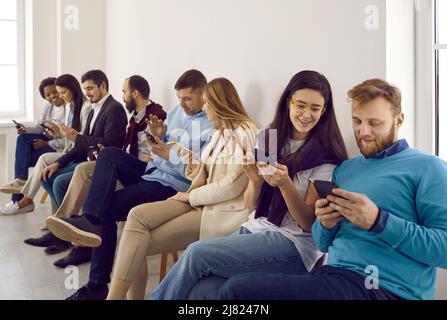 Gruppe glücklicher junger Menschen, die in Reihe sitzen und Social-Media-Apps auf Mobiltelefonen nutzen Stockfoto