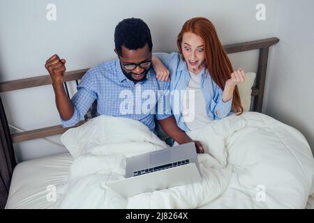 Ingwer rothaarige europäische Frau und hübscher afro afrikanischer Mann umarmen sich zusammen im Schlafzimmer zu Hause gemütlich Wohnung liegen und Nachrichten auf Laptop beobachten Stockfoto