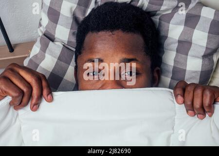 Junger, hübscher afro-amerikanischer Mann, versteckt unter einer weißen Decke in kuscheligem Baumwollkissen Stockfoto