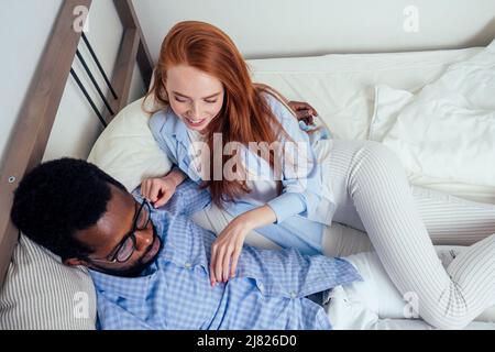 Ingwer Haar europäische weibliche und schöne afro afrikanischen Mann zusammen umarmt liegen im Schlafzimmer zu Hause gemütliche Wohnung Stockfoto