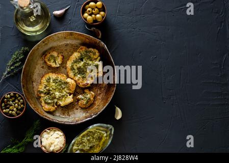 Blumenkohlesteak mit Gewürzen liegt in einer Pfanne. Olivenöl, Chimichurri-Sauce, Kräuter, verschiedene Gewürze nebeneinander. Dunkler Hintergrund. Für Text platzieren. Stockfoto