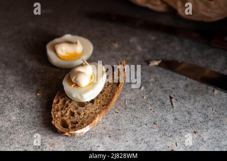 Hälften Hühnereier mit Mayonnaise und braunem Brot auf einem dunklen Küchentisch. Stockfoto