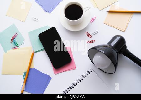 Draufsicht auf eine Tasse Kaffee in der Nähe von Schreibwaren, Lampe und Smartphone mit leerem Bildschirm auf Weiß Stockfoto